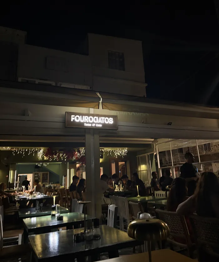 Η πρόσοψη του Fourogatos το βράδυ: η φωτεινή ταμπέλα πάνω από την είσοδο και τα τραπέζια μπροστά.