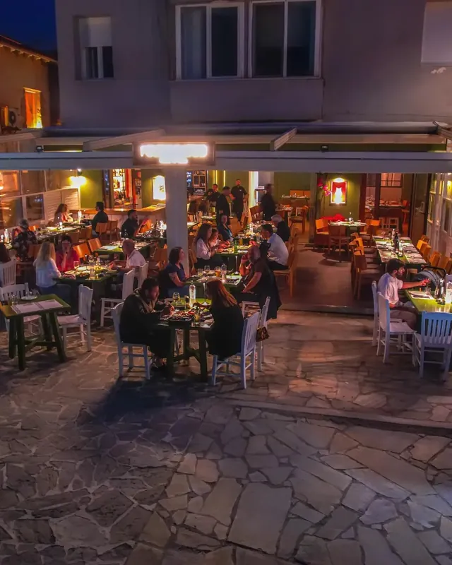 Η πέτρινη αυλή το βράδυ, με γεμάτα τραπέζια μπροστά από την πέργκολα.